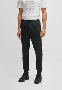 Svarta sportbyxor med en texturerad yta, vita sidostreck, elastisk midja och fickor med dragkedja. Bärs med en vit t-shirt med grafiskt tryck.