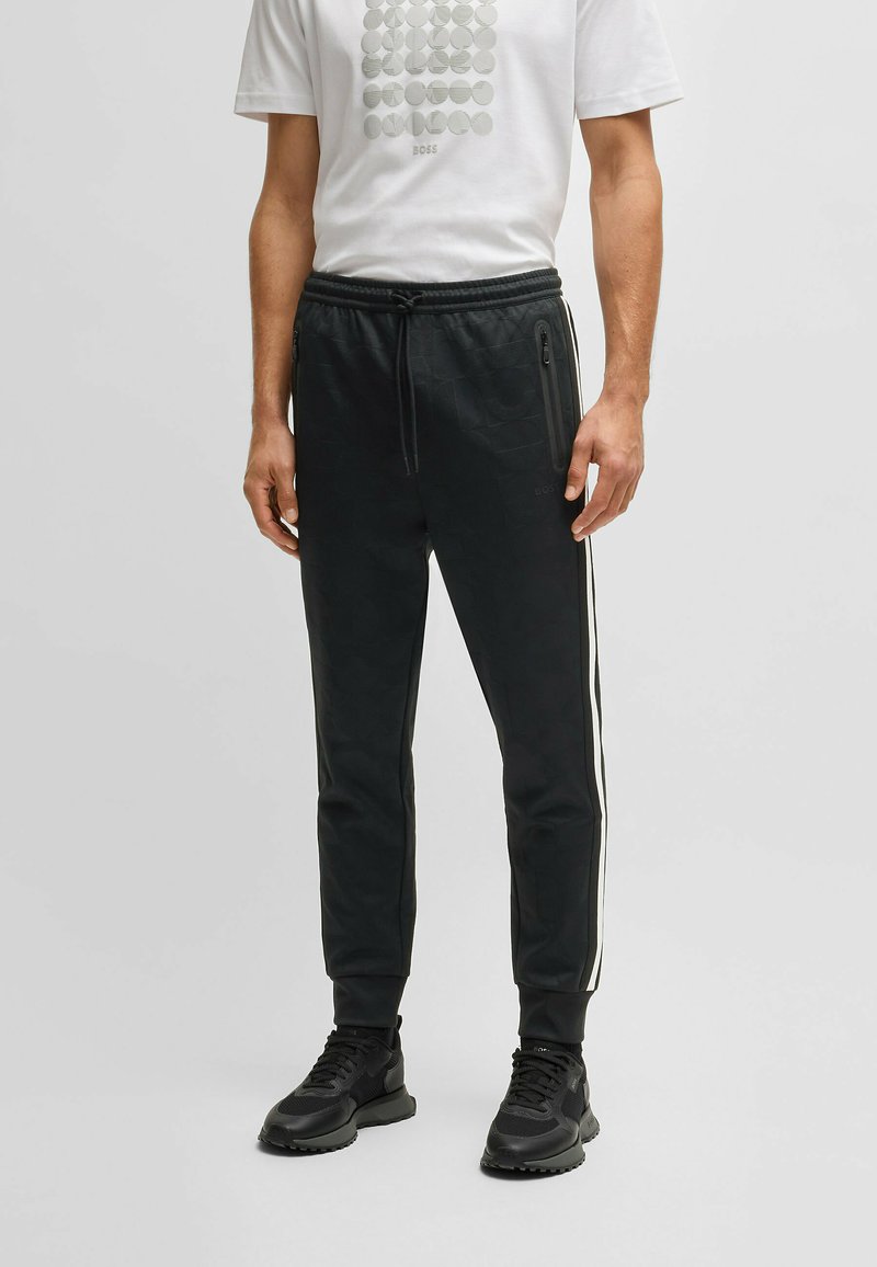 Svarta sportbyxor med en texturerad yta, vita sidostreck, elastisk midja och fickor med dragkedja. Bärs med en vit t-shirt med grafiskt tryck.