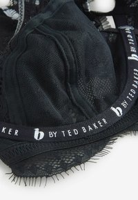 Čierna čipkovaná podprsenka s dizajnom z oka, vybavená elastickou páskou s logom značky. Výrazné detaily zahŕňajú zvlnené okraje a jemné variácie textúry.