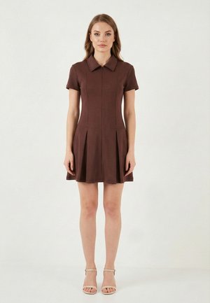 Femme portant une robe marron à manches courtes avec col et plis, debout les bras le long du corps, chaussée de talons beiges à bride autour de la cheville, sur un fond blanc.