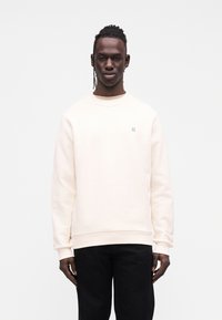 Φούτερ κρεμ χρώματος με μακριά μανίκια, στρογγυλή λαιμόκοψη και κεντημένη λεπτομέρεια στο στήθος· σε συνδυασμό με μαύρο παντελόνι.