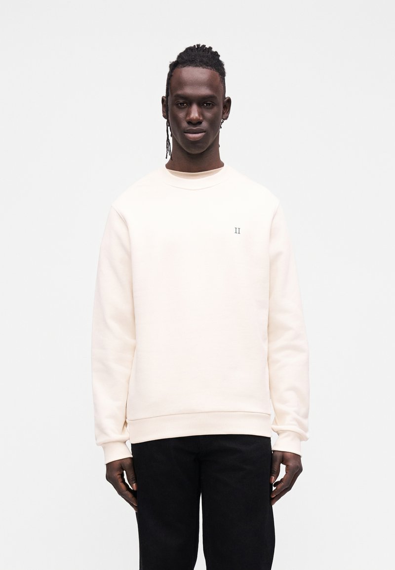 Φούτερ κρεμ χρώματος με μακριά μανίκια, στρογγυλή λαιμόκοψη και κεντημένη λεπτομέρεια στο στήθος· σε συνδυασμό με μαύρο παντελόνι.