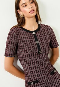 Kurzärmeliges Kleid mit schwarzem und pinkem geometrischem Muster. Verfügt über einen runden Ausschnitt, durchgehende Knopfleiste und zwei Taschen mit Knopfakzenten.