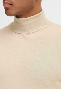 Pulover bej cu guler înalt, având o textură dreptunghiulară și un design mulat. Gulerul este strâns, cu o finisare fără cusături în jurul gulerului.