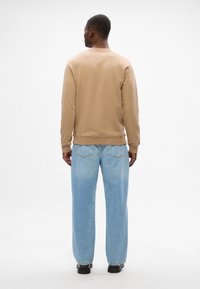 Felpa beige in cotone con maniche lunghe e polsini a coste, abbinata a jeans in denim blu chiaro a vestibilità ampia. Texture liscia e design semplice.