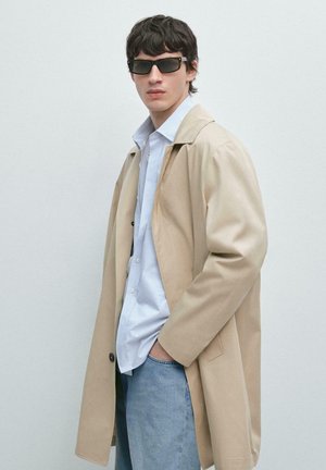 Mladý muž nosící černé sluneční brýle, béžový kabát, světle modrou košili a modré džíny, stojící s jednou rukou v kapse před jednobarevným pozadím.