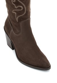 Bota vaquera de ante marrón con punta afilada, tacón de madera apilado y detallado de costura de inspiración occidental en contraste.