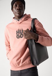 Sudadera con capucha en rosa suave con impresión negra "HUGO", combinada con un bolso de cuero negro con forma rectangular y superficie texturizada.
