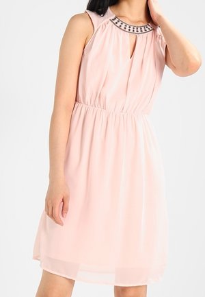 Robe sans manches rose pâle longueur genou avec taille froncée et encolure ornée de bijoux portée par une femme aux cheveux foncés.
