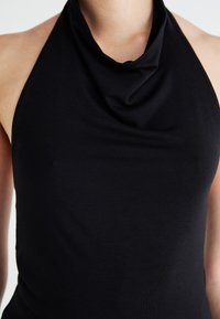 Μαύρο τοπ με λαιμόκοψη halter σε μαλακό, ελαστικό ύφασμα που διαθέτει draped λαιμό. Η υφή είναι λεία χωρίς ορατά μοτίβα ή λεπτομέρειες.