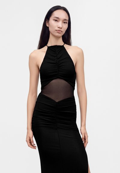Femme aux longs cheveux foncés portant une robe noire sans manches avec des détails froncés et un panneau transparent au niveau du milieu, debout devant un fond uni.