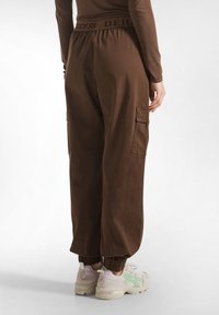 Bruine cargobroek met elastische boorden en tailleband, met een "GODDESS" label. Inclusief zijzakken voor extra functionaliteit.