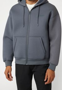 Sudadera gris con cremallera en tela suave, que cuenta con capucha, bolsillos frontales y acentos de costura blanca. Diseño simplista con detalle de logo.