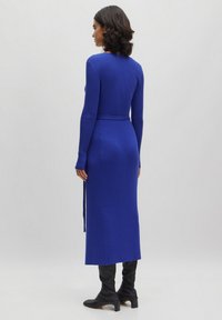Rippstrickkleid in elektrisiertem Blau mit taillierter Silhouette, langen Ärmeln, einem Taillengürtel und knielanger Länge. Getragen mit knielangen schwarzen Stiefeln.