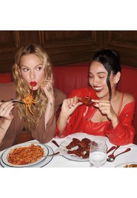 Zwei Frauen essen an einem Tisch. Die eine hält Spaghetti mit Tomatensauce; die andere genießt Hähnchenflügel. Ein Glas Sprudelwasser steht auf dem Tisch.