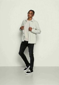 Jovem com cabelo trançado, vestindo uma jaqueta cinza clara sobre uma camisa bege, jeans skinny pretos e sapatilhas pretas com solas brancas.