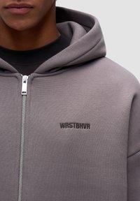Felpa grigia con zip realizzata in tessuto testurizzato, con un ampio cappuccio e il logo ricamato "WRSTBHV" sul petto. Dettagli in zip argentati visibili.