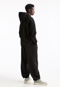 Črn oversized hoodie z vrvico na kapuci, v kombinaciji s širokimi črnimi trenirkami in sivimi supergami. Enostavna, gladka tekstura tkanine.