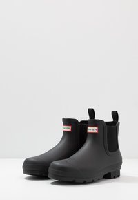Black rubber ankle boots with a smooth finish, elastic side panels, and distinctive branding on the front. Textured rubber soles for traction.