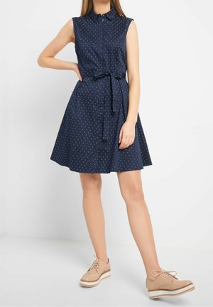 Granatowa sukienka bez rękawów z drobnym białym wzorem, z paskiem w talii, kołnierzykiem i zapinana z przodu na guziki, noszona z beżowymi platformowymi oksfordkami.