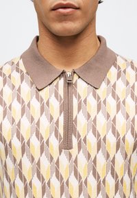 Κοντινό πλάνο ενός ατόμου που φοράει πουλόβερ με γεωμετρικά σχέδια σε καφέ, κίτρινο και λευκό, με καφέ γιακά και φερμουάρ μπροστά.