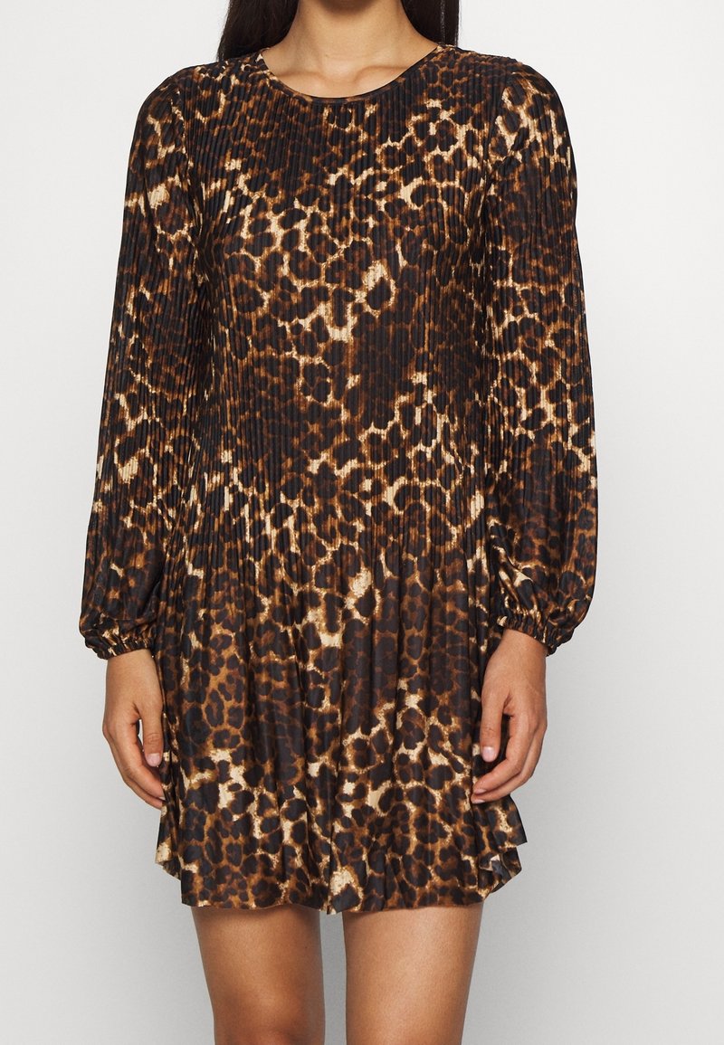 Femme portant une robe plissée à manches longues avec un imprimé léopard marron et noir, debout devant un fond clair uni.