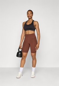 Crni sportski grudnjak, smešnate visoke kratke hlače, drži crnu kettlebell. Bele čarape i svetla sportska obuća. Minimalistička pozadina.