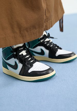 Person iført brune bukser og hvide, sorte og teal høje sneakers, der står på en lyseblå overflade.