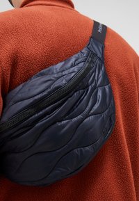 Quilted navy nylon sling bag with a curved shape and zip closure, worn over an orange fleece garment. Textured with a wave pattern.