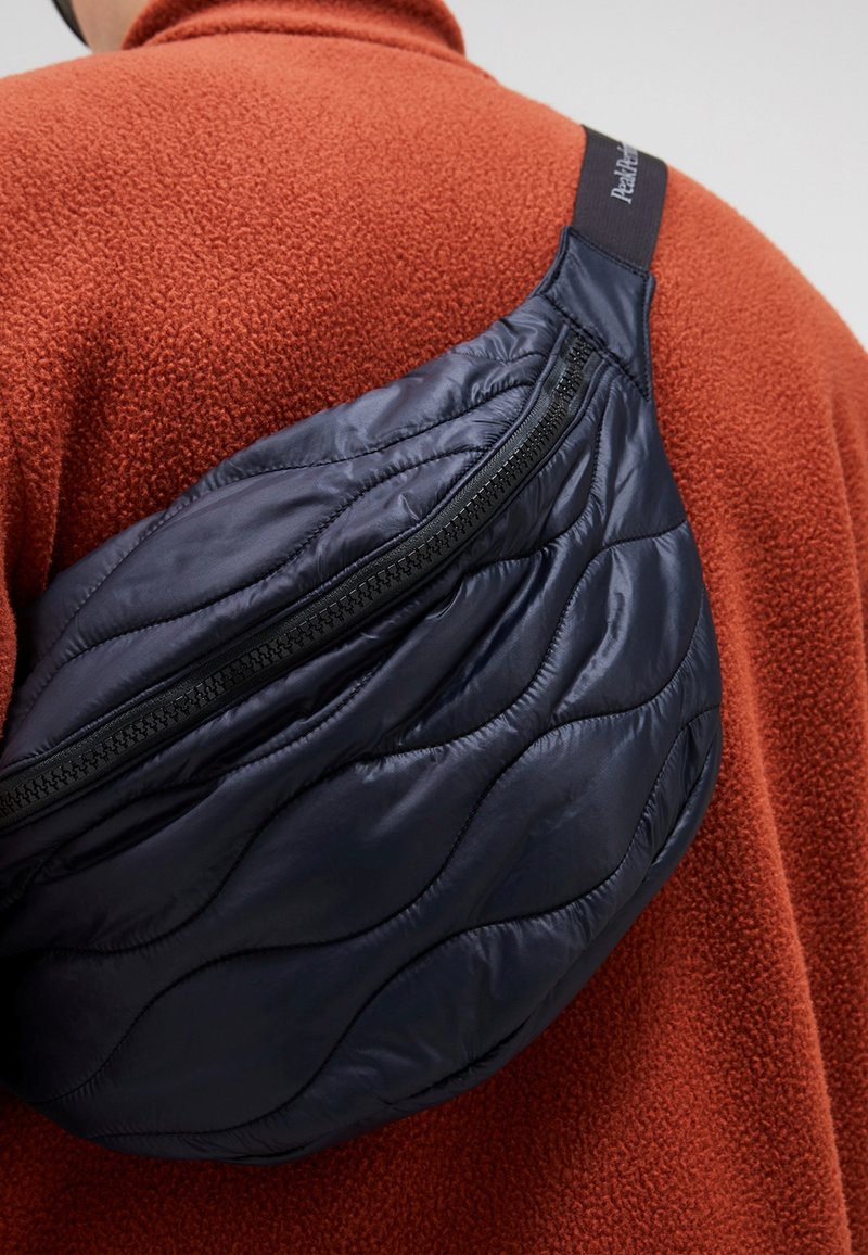 Quilted navy nylon sling bag with a curved shape and zip closure, worn over an orange fleece garment. Textured with a wave pattern.