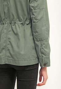 Personne portant une veste verte avec une taille élastique et des poignets à boutons, associée à un jean noir, montrant une vue de dos et de côté du torse et du bras.