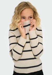 Młoda osoba z falującymi blond włosami, ubrana w kremowy sweter z prążkami w kolorze czarnym, częściowo zasłaniająca twarz kołnierzem swetra.