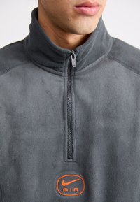 Γκρι φούτερ με φερμουάρ μέχρι τον λαιμό, σε στυλ quarter-zip, που διαθέτει ένα μικρό πορτοκαλί λογότυπο Nike AIR στο στήθος και φερμουάρ στην περιοχή του λαιμού. Μαλακή υφή και εφαρμοστό σχέδιο.