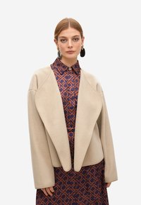 Mujer con el cabello recogido, que lleva grandes pendientes negros y dorados, una chaqueta beige de corte oversized y un vestido a rayas en tonos azul marino y naranja con cuello.