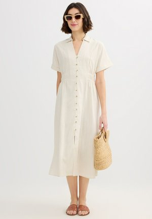 Mujer con vestido midi beige abotonado, sandalias color canela, gafas de sol y sosteniendo un bolso de paja tejido frente a un fondo blanco.