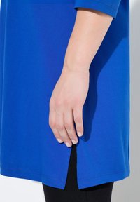 Blaue Baumwolltunika mit geradem Schnitt, versehen mit einem seitlichen Schlitz und glatter Textur, kombiniert mit schwarzen Leggings. Die Hand ruht auf dem Rand der Tunika.