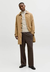 Homme debout, portant un trench-coat beige, un pull crème à demi-fermeture éclair, un pantalon ample à carreaux marron et des mocassins noirs, sur fond blanc uni.