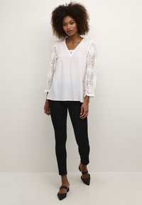 Weiße Bluse mit kunstvollen Spitzenärmeln, V-Ausschnitt und lässiger Passform, kombiniert mit schwarzen Skinny Jeans und schwarzen Absätzen.