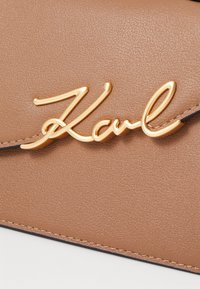 Tan faux skinnveske med hevet gull "Karl"-logo. Glatt tekstur med en lett glans og kontrasterende mørk søm.