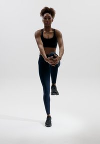 Svart sport-bh, marinblå leggings och svarta sneakers; individen sträcker sig genom att höja ett knä och greppa det med båda händerna.