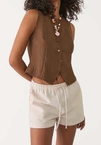 Brązowy bezrękawnik z guzikami o teksturowanym wykończeniu, w zestawieniu z jasnobeżowymi szortami na sznurek. Uzupełniony kolorowym naszyjnikiem z koralików.