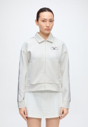 Femme aux cheveux foncés attachés, portant une veste zippée gris clair avec des rayures noires sur les manches et une courte jupe blanche.