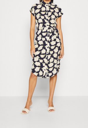 Woman wearing a navy shirt dress with white floral print, a waist tie, and beige braided heeled sandals, standing against a plain background.