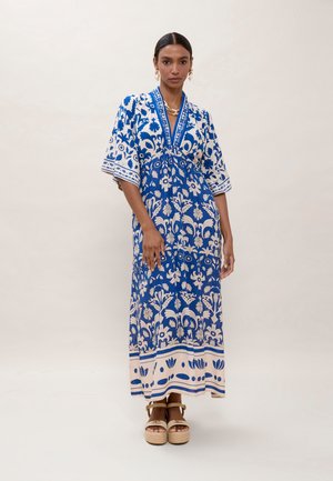 Mujer vestida con un vestido maxi de patrón floral en azul y beige, con mangas hasta el codo y sandalias de plataforma beige, de pie frente a un fondo liso.