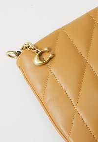 Coin de sac à main matelassé en cuir couleur fauve avec chaîne en métal doré et détail de charme en forme de fer à cheval sur fond blanc.