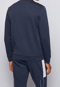 Granatowa bluza z okrągłym dekoltem, z prążkowanymi mankietami i suwakiem, wykonana z gładkiej tkaniny. Charakteryzuje się minimalistycznym designem bez widocznych logo i wzorów.