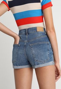 Jeansshorts i medelblått med fransar i fållen och en ljusblå uppvikning. Har två bakfickor och en rektangulär beige etikett på midjebandet.