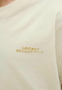 Kremkrāsas kokvilnas džemperis ar apaļu kakla izgriezumu un ribinātām piedurknēm, ar zelta drukātu logo "LOCAL* COLLECTIVE" priekšpusē.