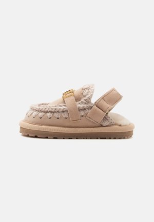 Pantofole beige aperte sul retro con fodera in lana, dettagli in maglia intrecciata, cinturino regolabile con fibbia dorata e suola in gomma, vista laterale.