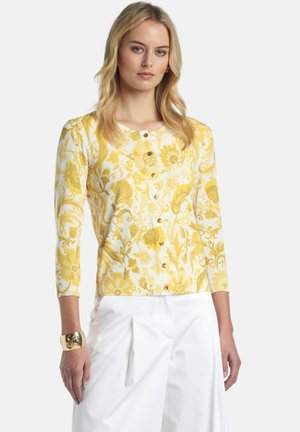 Vrouw die een gele en witte bloemrijke gebloemde cardigan met knopen en driekwart mouwen draagt, gecombineerd met witte geplooide broek en een gouden armband als accessoire.